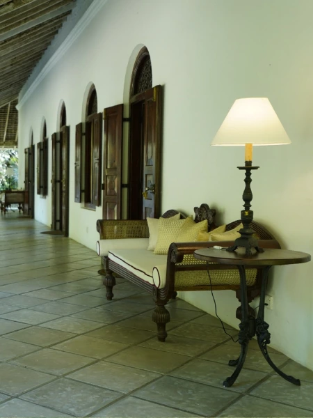 Colonial-style veranda at Adigar’s Manor with wooden shuttered windows, a vintage settee, and a classic table lamp, evoking timeless charm and relaxation.