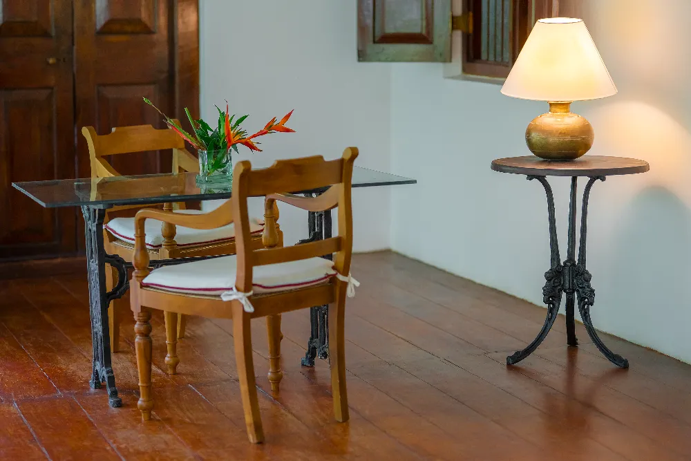 Elegant colonial-style dining nook with wooden chairs, glass-top table, tropical flowers, and warm ambient table lamp