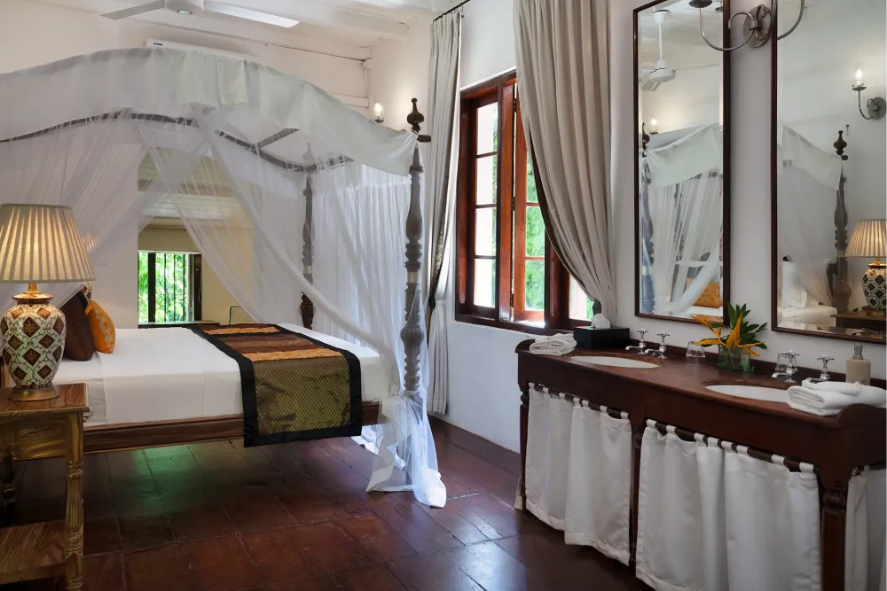 Elegant Indian Admiral Room featuring a four-poster canopy bed, wooden vanity with double sinks, tall mirrors, and warm colonial-style interiors