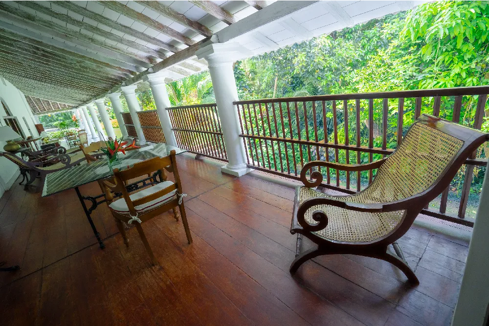 Wide colonial-style balcony outside the Indian Admiral Room featuring wooden lounge seating, dining area, white pillars, and lush tropical forest views