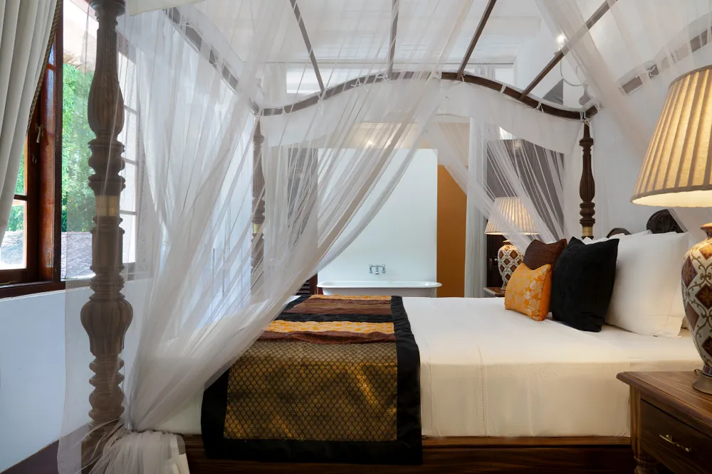 Luxurious Indian Admiral Room with a four-poster canopy bed, flowing white drapes, warm bedside lighting, and a freestanding bathtub in the background