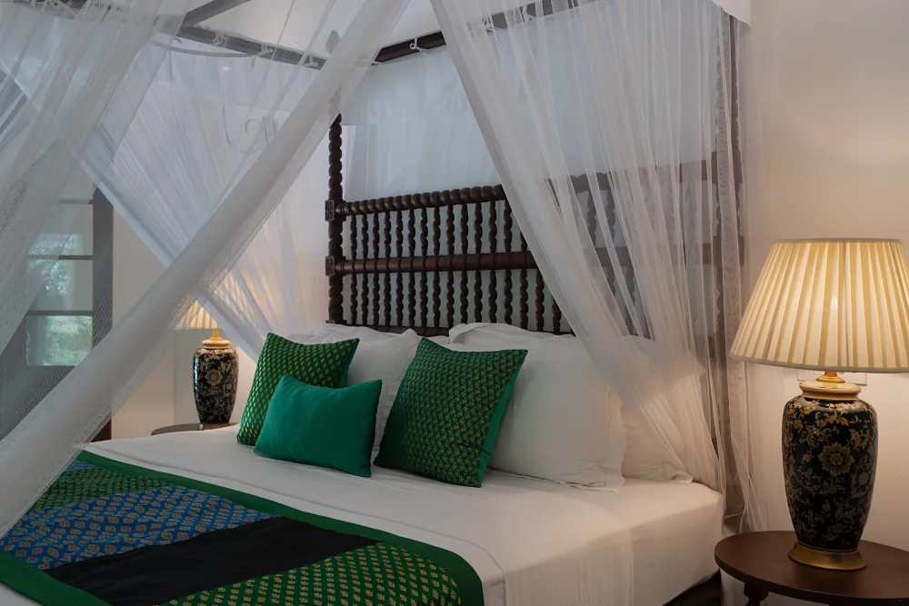 Peacock Royal Room featuring a luxurious four-poster canopy bed, elegant sheer drapes, artisan ceramic lamps, and rich emerald accents in a serene colonial-style setting