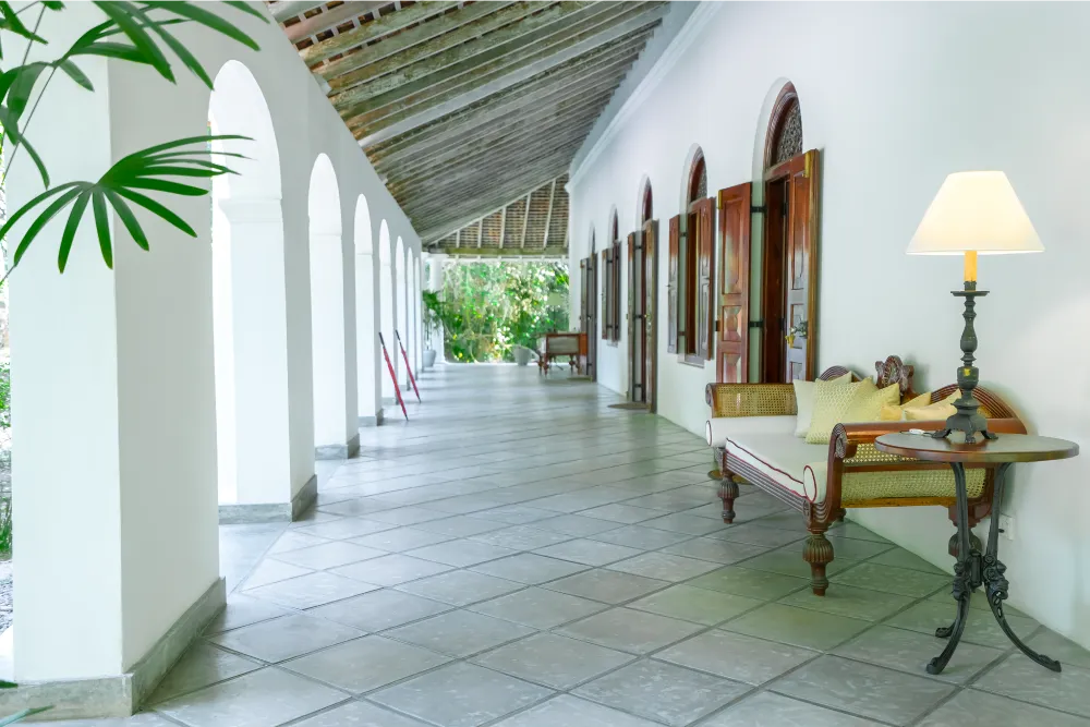 Elegant colonial-style corridor with white archways, polished tiled flooring, antique wooden seating, and warm ambient lighting overlooking lush tropical greenery