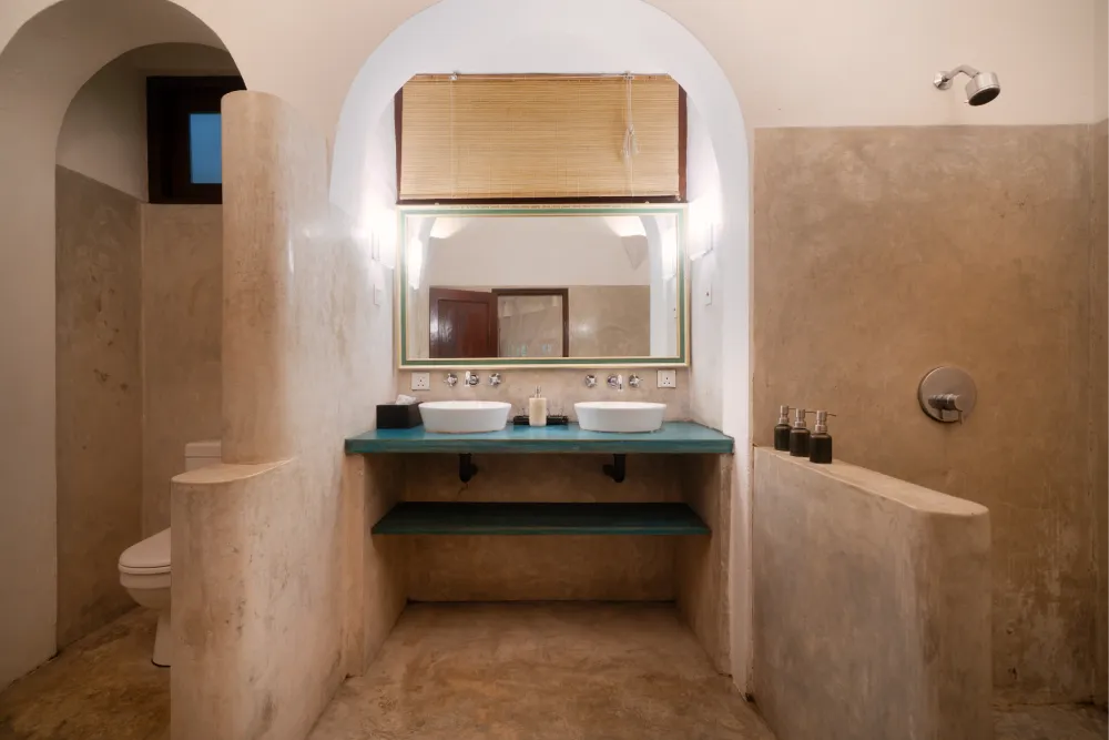 Spacious Pioneer Room bathroom with twin vessel sinks, natural stone walls, walk-in shower, and elegant arched mirror