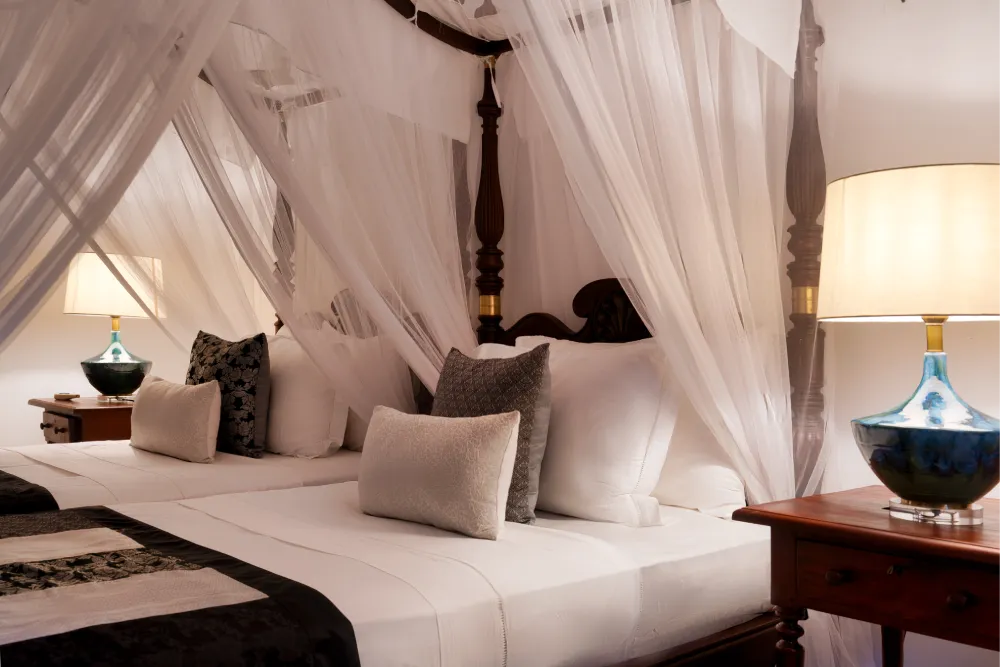 Elegant Pioneer Room with four-poster canopy bed, white mosquito netting, soft neutral cushions, and warm bedside lighting