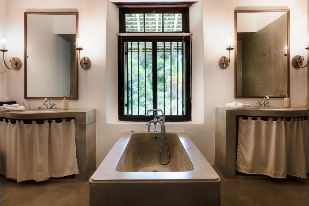 Redspot Duke Room bathroom featuring a luxurious stone soaking tub, twin minimalist vanities, elegant wall sconces, and serene colonial-inspired interiors with tropical views