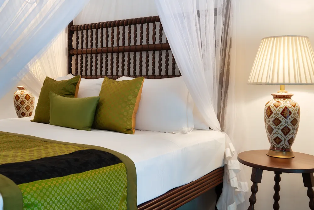 Redspot Duke Room with elegant carved wooden bed, flowing canopy drapes, warm bedside lighting, and sophisticated colonial-inspired décor