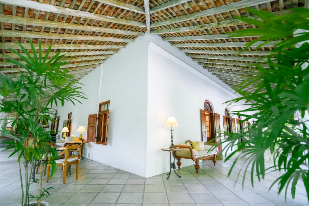 Spacious colonial-style veranda with high timber ceilings, elegant lounge seating, warm ambient lighting, and lush tropical greenery framing the walkway