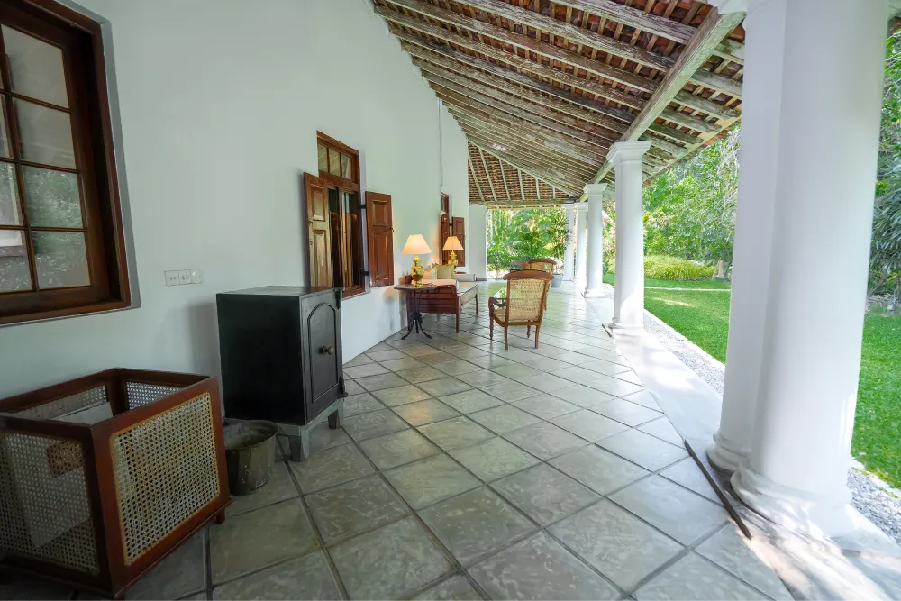 Elegant colonial-style veranda with white pillars, tiled flooring, vintage wooden furniture, and peaceful tropical garden surroundings
